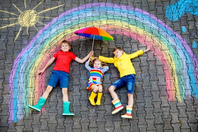 Children wearing colourful cloths laying on paving under a chalk rainbow