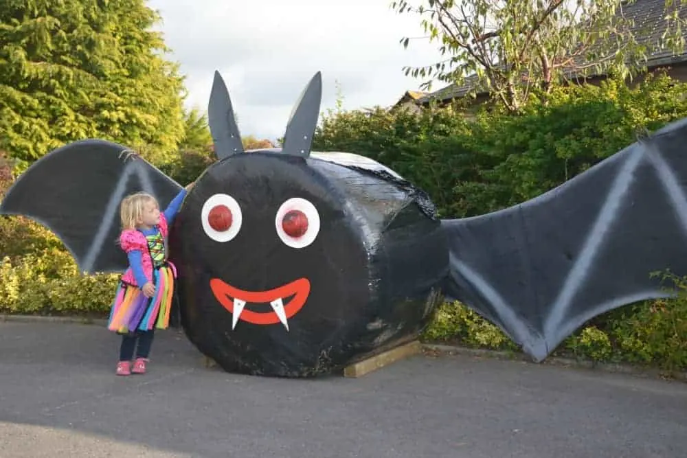 Child in witches costume stood with giant bat made from a covered hay bale