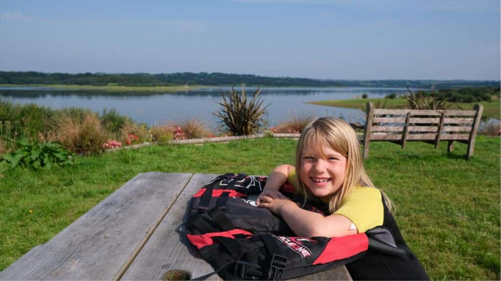 Child sitting at picnic bench by lake with buoyancy aid