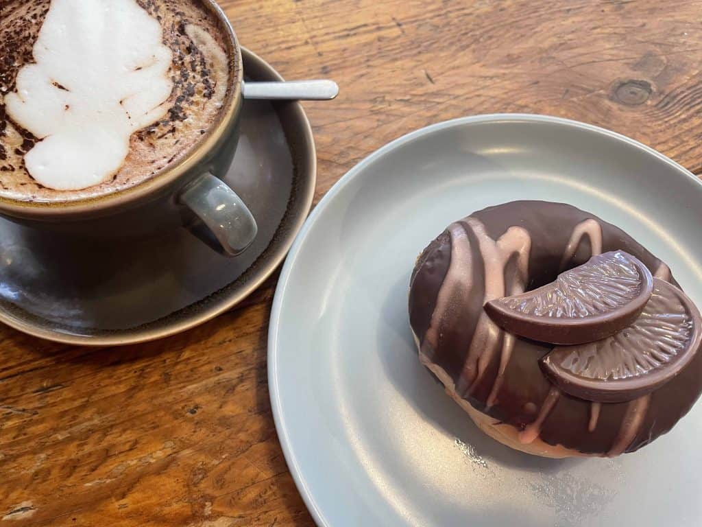 Donut with slices of Terry's Chocolate Orange decorating the top next to a frothy coffee in cup and saucer