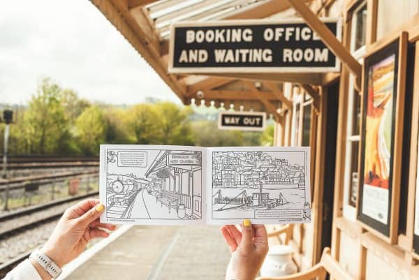 Colouring book page with an illustration of Totnes Station being held up in front of the same view