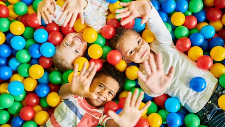Three children wave at camera while laid in ball pit