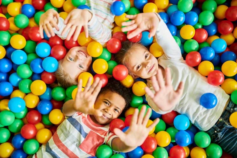 Three children wave at camera while laid in ball pit