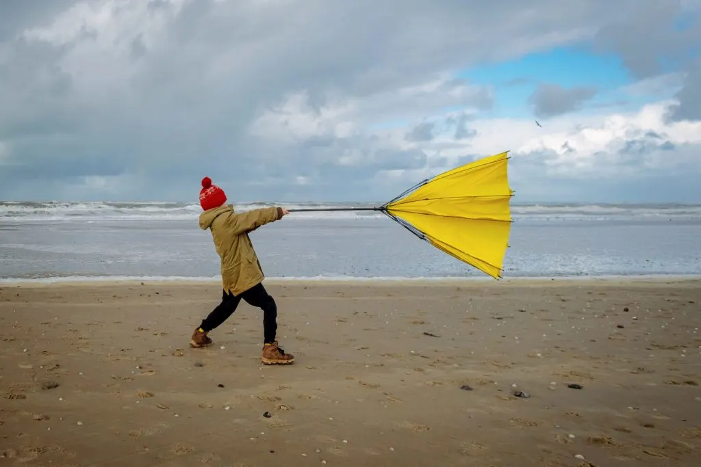 Young boy struggling with large yellow umbrella on beach in stormy weather.