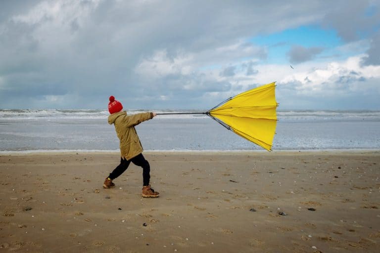 Young boy struggling with large yellow umbrella on beach in stormy weather.
