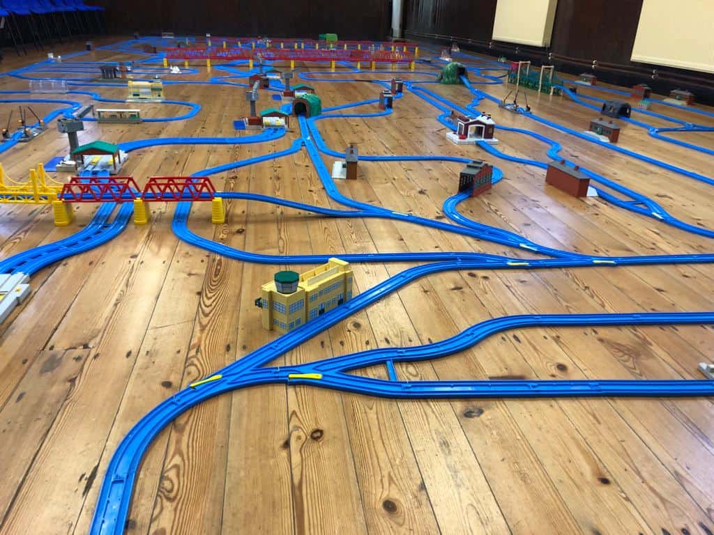 Large blue toy train track laid out on a wooden floor with stations and other railway features
