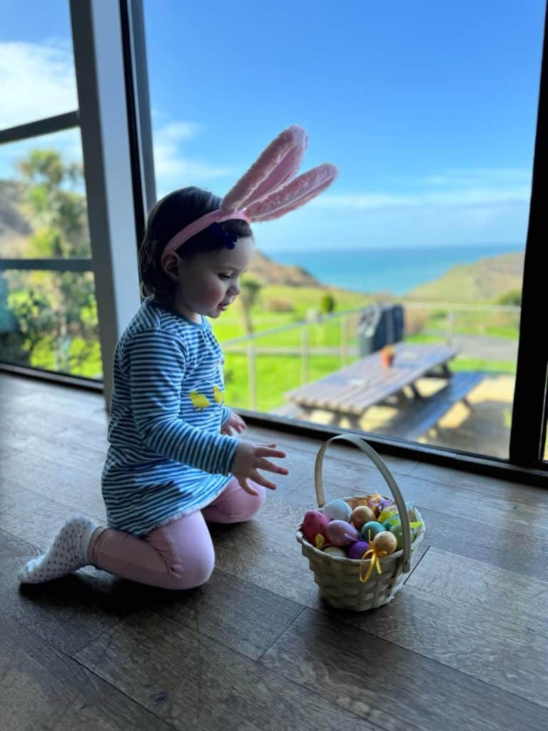 Young girl wearing bunny ears kneeling in front of a basket of colourful eggs with a a view of the South Devon landscape behind her
