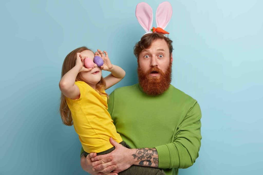 Scared emotional red haired man with bunny ears carries little cheerful girl who holds two colored eggs on eyes, celebrate Easter together, isolated over blue wall. Dad with kid prepare for holiday