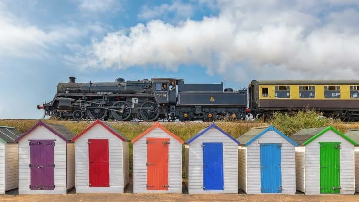 Dartmouth Steam Railway steam engine traveling along the line between Kingswear and Paignton. There are colourful beach huts at Goodrington Sands in the foreground