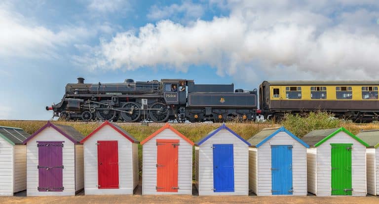 Dartmouth Steam Railway steam engine traveling along the line between Kingswear and Paignton. There are colourful beach huts at Goodrington Sands in the foreground