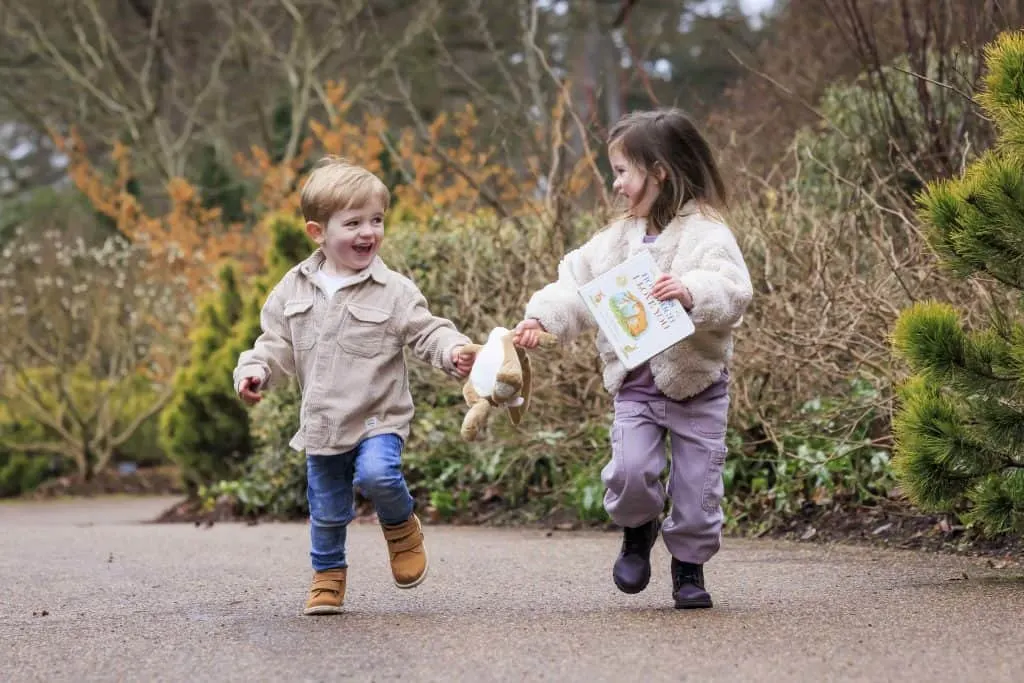 Two children run along a garden path with a copy of the children's book Guess How Much I Love You
