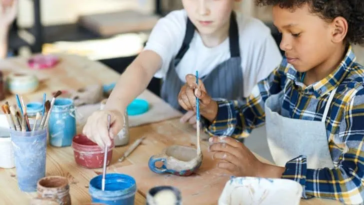 Two children sitting by table and painting self-made clay items at lesson