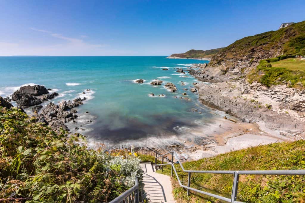A view out across Combesgate Beach in Woolacombe. It is a sunny day and the sea is blue. The beach is a narrow arch lined with rock pools
