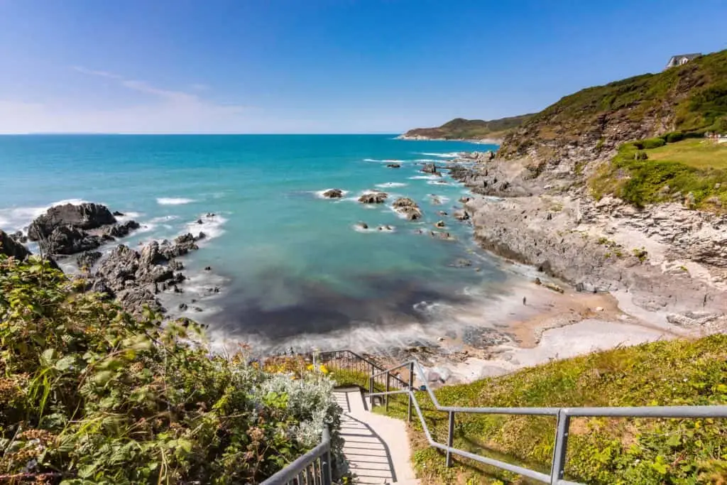 A view out across Combesgate Beach in Woolacombe. It is a sunny day and the sea is blue. The beach is a narrow arch lined with rock pools
