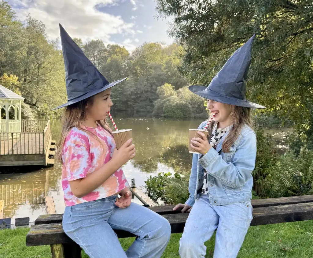 Two girls wearing witches hats sit beside the lake with milkshakes at Canonteign Falls