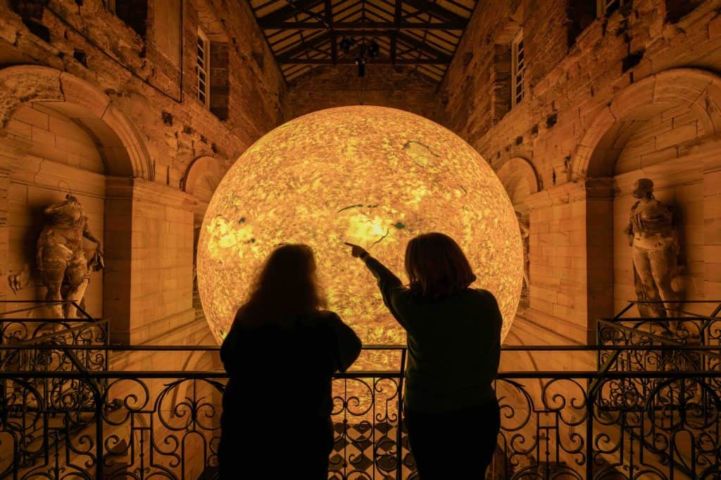 Two people stand in front of the Helios installation in a large gothic design room
