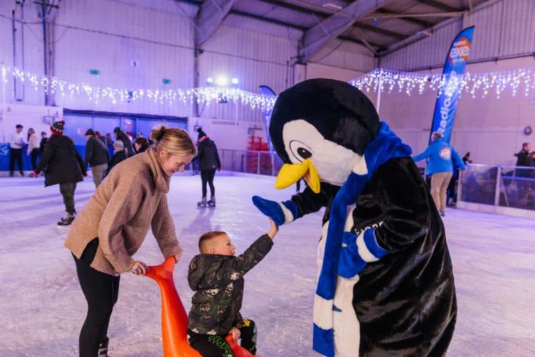 Child on skating aid being pushed by parent. A penguin mascot is offering the child a high five