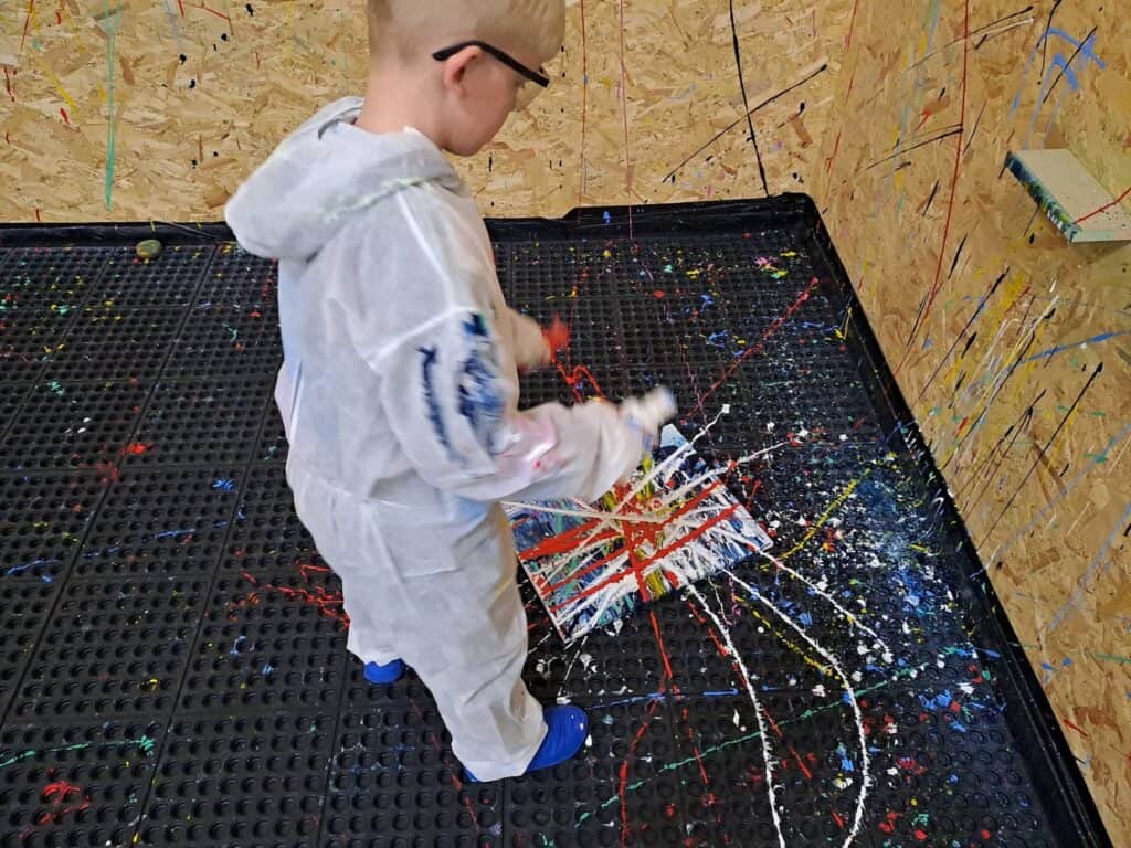 Young boy in a painting overall splattering multi-coloured paint at a wall