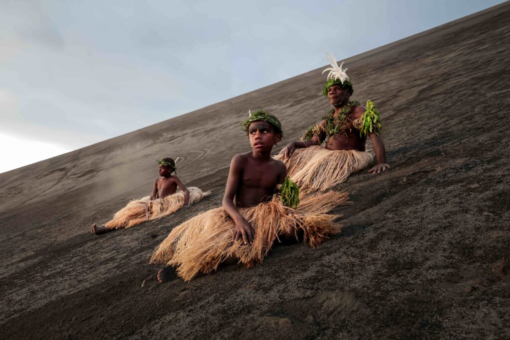 Three people in national dress sit on the baked ground of the side of a volcano